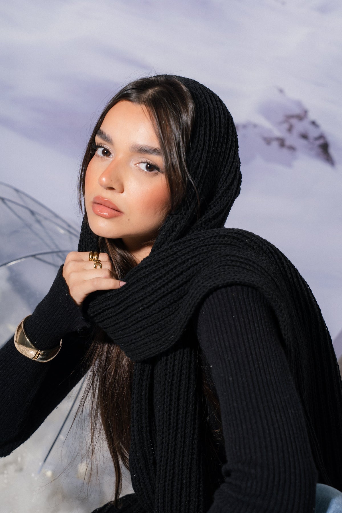 A woman wearing a black ribbed knit hooded sweater and matching scarf, posed against a snowy mountain backdrop.