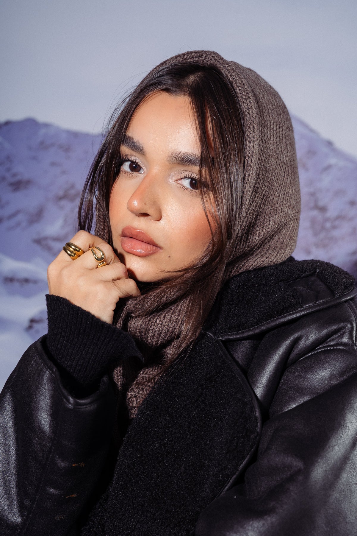 A woman wearing a black faux-leather jacket, brown knit hood, and brown scarf, posing in front of a snowy mountain backdrop.