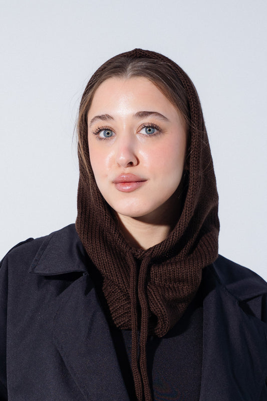 A woman wearing a dark brown ribbed knit hooded neck warmer or cowl under a black coat, photographed against a plain light gray studio background.