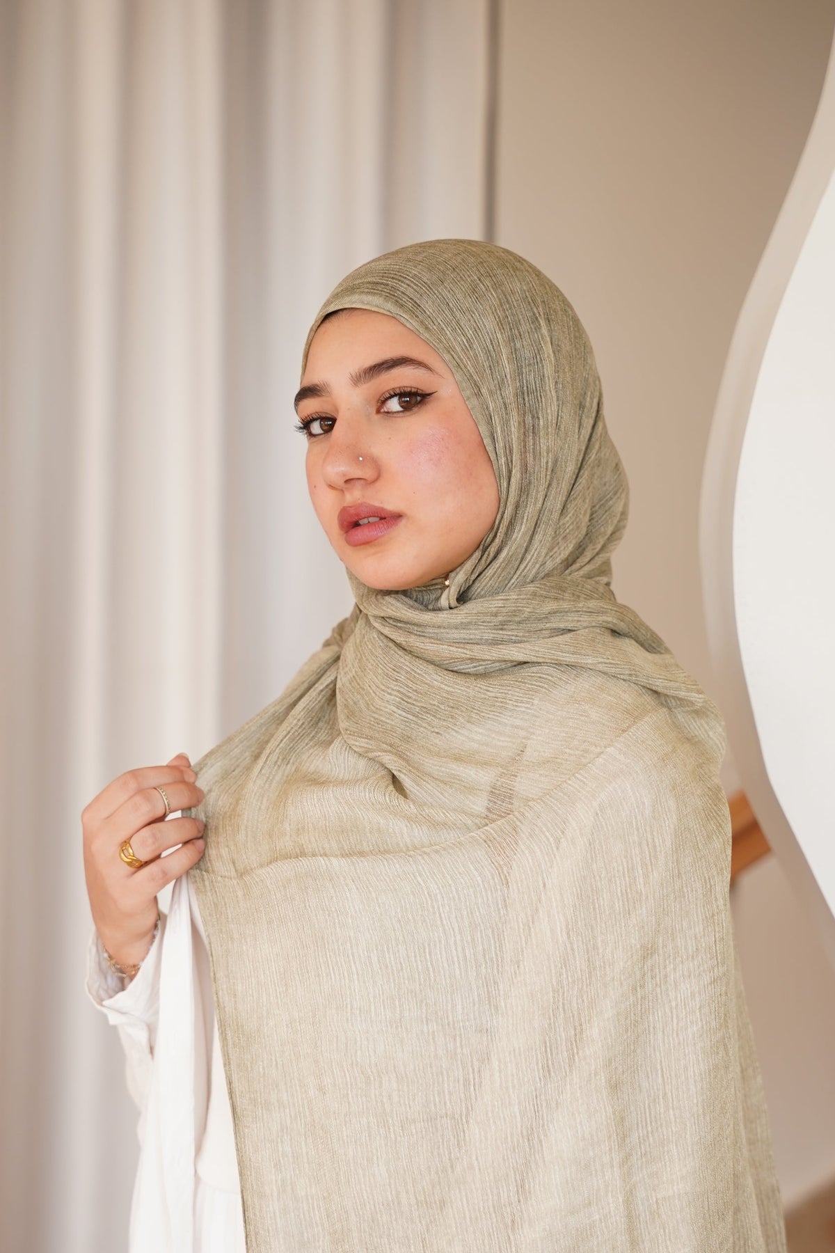A woman wearing a light beige textured hijab and a white dress, standing in a modern interior with wavy white walls and a staircase.