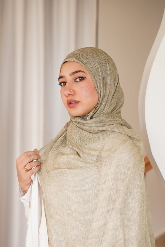 A woman wearing a light beige textured hijab and a white dress, standing in a modern interior with wavy white walls and a staircase.