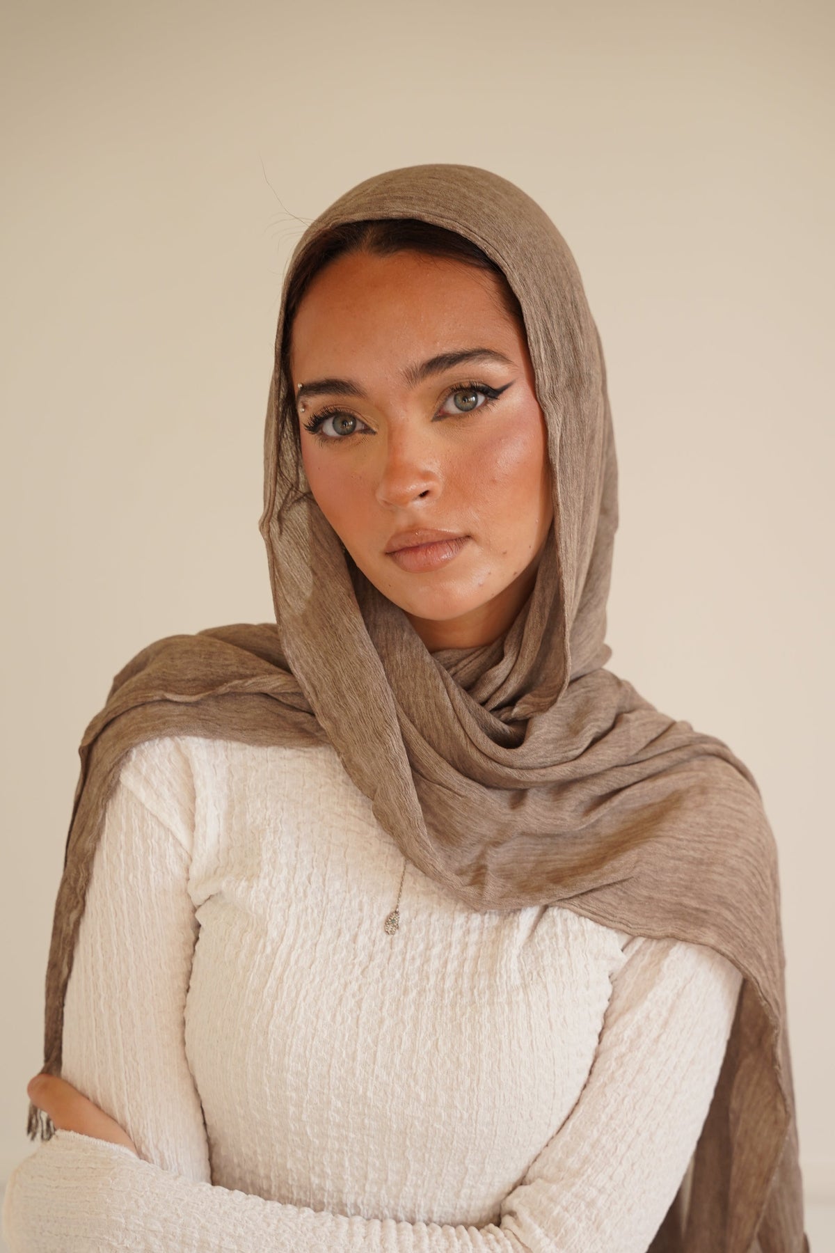 A woman wearing a taupe brown hijab and a white textured long-sleeve top, posing against a neutral cream-colored wall.