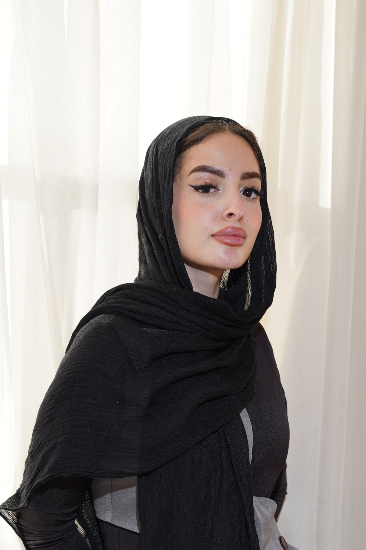 A woman wearing a black hijab and a sheer black shawl draped over her shoulders, posed in front of white sheer curtains.